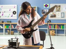 High-resolution realistic photograph of a student holding or playing a 3D-printed electric guitar in a university lab or music room, photographed in a real environment, matching the look of existing press images about the same project. Avoid futuristic or stylized elements.